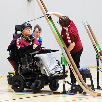 Sähköpyörätuolia käyttävä Vivi Virtanen ja peliavustaja asettelevat pelipalloa pelikouruun paraboccia-ottelussa. Vivi käyttää kypäräänsä kiinnitettyä vipua pallon asetteluun.